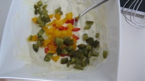 In einer weißen Schüssel wird ein sahniger Eiersalat mit bunten Paprika und Gewürzgurken angerichtet. Ein Schneebesen liegt daneben.