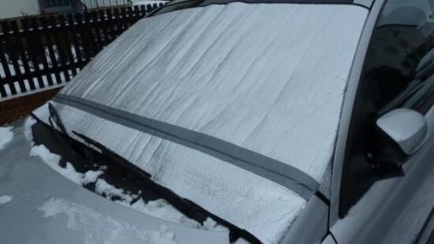 Eine Abdeckung schützt die Frontscheibe eines Autos vor Schnee, während es auf einem verschneiten Parkplatz steht.