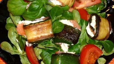 Auf einem schwarzen Teller liegt Feldsalat mit gebratenen, gefüllten Zucchinirollen und frischen Tomatenscheiben.