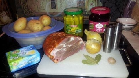 Ein Schweinebraten und Gewürze liegen auf einem Schneidebrett, daneben Kartoffeln, Gewürzgurken und Rote Bete.