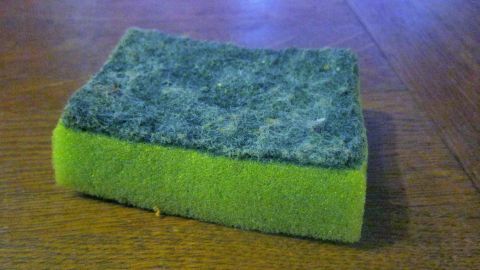 Ein gebrauchter grüner Spülschwamm liegt unbewegt auf einem Holztisch, zeigt Abnutzungsspuren auf der dunklen Scheuerfläche.