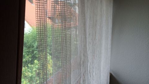 Transparente Gardinen hängen vor einem Fenster und lassen Licht herein, während die Aussicht auf einen Garten sichtbar bleibt.