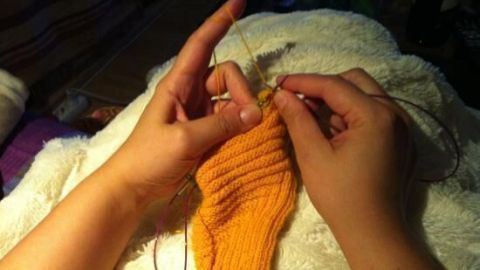 Helle Hände halten eine gelbe Socke mit einer Rundstricknadel, während sie bequem auf einem weißen Tuch sitzt.