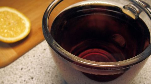Ein Glas schwarzer Tee mit einem Ölfleck schwimmt auf der Oberfläche, während ein Zitronenschnitz daneben liegt.