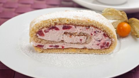 Eine Biskuitrolle mit Erdbeer-Sahne ist auf einem weißen Teller angerichtet, bestäubt mit Puderzucker und umgeben von einer bunten Tischdecke.