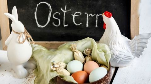 Ein weißer Hase und ein Huhn umgeben ein Holztablett mit bunten Eiern und Schokoladenfiguren, beschriftet mit "Ostern".