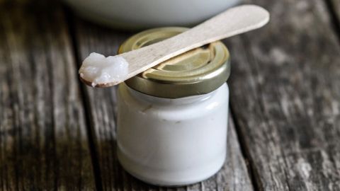 Kokosnussfett gilt als hervorragende Pflege für weiche Haut. Ein Glas mit Kokosnussfett steht auf einer rustikalen Holzoberfläche, daneben ein kleines Glas mit einem Holzlöffel, darauf eine Creme.