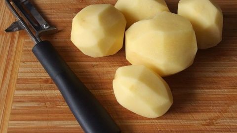 Kartoffeln liegen auf einem Holzbrett, einige geschält, daneben ein Schäler, bereit zum weiteren Schälen.