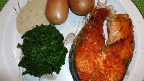 Ein knuspriges Lachssteak liegt neben Spinat, Kartoffeln und Dillsauce auf einem weißen Teller, alles auf grünem Tisch.