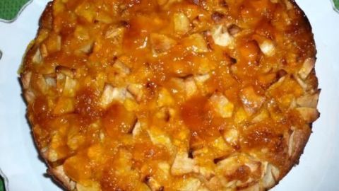 Ein Apfelkuchen mit Aprikosenguss, goldbraun gebacken, auf einem weißen Teller, steht auf einem grünen Untergrund.