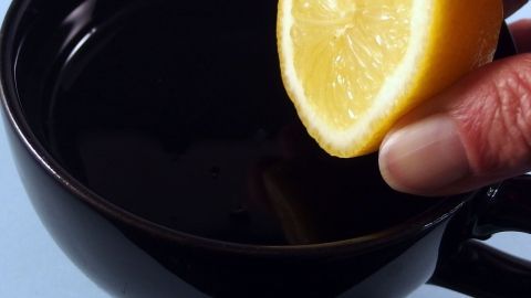 Eine Hand hält eine Zitronenscheibe über eine schwarze Tasse, während der dunkle Kaffee im Hintergrund sichtbar ist.