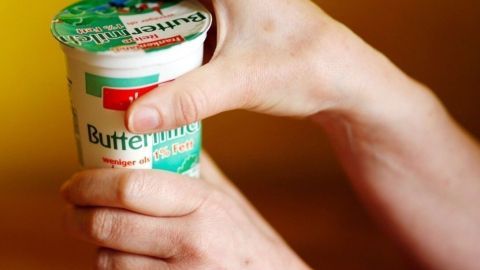 Zwei Hände drücken auf einen Becher Buttermilch, während der Hintergrund in warmen Gelbtönen gehalten ist.