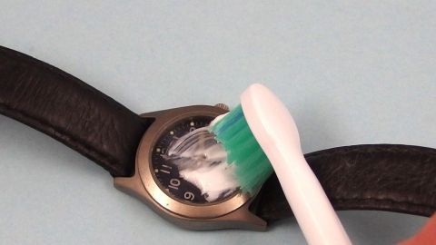 Eine Uhr wird mit einer Zahnbürste gereinigt, während sie auf einem blauen Hintergrund liegt, um Kratzer zu entfernen.