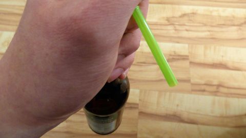Eine Hand hält einen grünen Strohhalm, der in ein Getränk eingetaucht ist, während der Hintergrund aus Holzboden besteht.