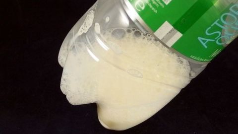 Kühlen Milchschaum in einer Selterflasche herstellen. Eine halbvolle Plastikflasche schüttet schaumige Milch aus, die auf einem dunklen Untergrund landet.