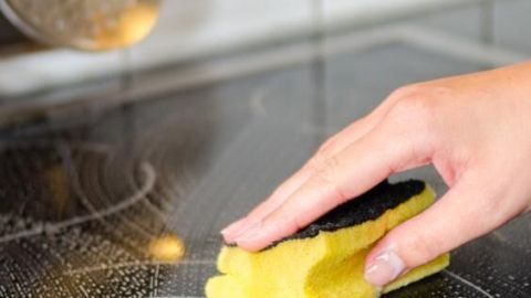 Eine Hand reinigt ein Ceranfeld mit einem gelben Schwamm in einer Küche, umgeben von weißen Fliesen und Küchenutensilien.