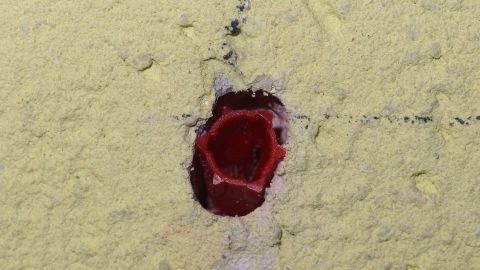 Ein roter Dübel steckt fest in einer gelben Wand, umgeben von Staub und kleinen Partikeln, was eine feste Verbindung zeigt.