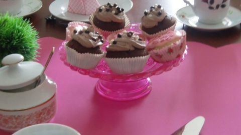 Auf einer rosa Tablett befinden sich schokoladige Muffins mit Sahne und Schokoladenstückchen, umgeben von zierlichen Tassen.