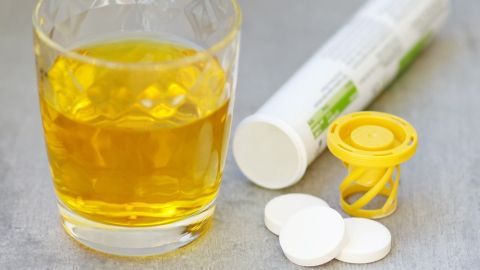 Brausetabletten liegen neben einem Glas mit gelbem Getränk; im Hintergrund eine geöffnete Verpackung auf grauem Tisch.