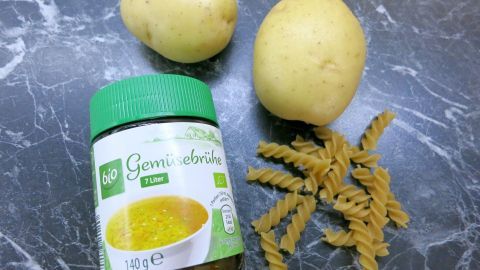 Mithilfe von etwas Brühe lässt sich das Überkochen beim Nudeln kochen vermeiden. Zwei kleine Kartoffeln liegen neben einer Packung Nudeln und einem Glas Brühe auf einer dunklen, marmorierten Oberfläche.