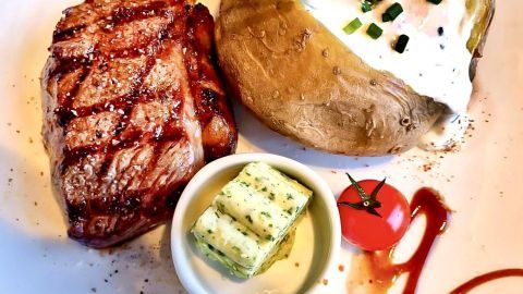 Ein saftiges Grillsteak liegt auf einem Teller neben einer gebackenen Kartoffel mit Sauce und Kräuterbutter.