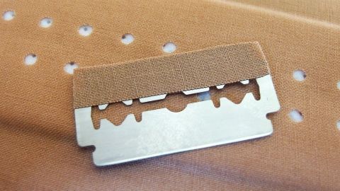 Ein Rasierklingen mit Klebeband zum Verletzungsfreien Basteln liegt auf einem Untergrund mit Löchern in brauner Farbe.