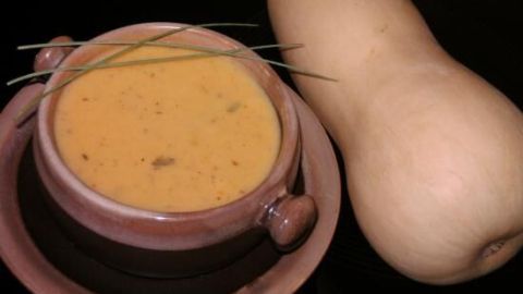 Eine Schüssel mit Kürbiscremesuppe steht neben einem Butternut-Kürbis auf einem schwarzen Tisch, dekoriert mit Schnittlauch.