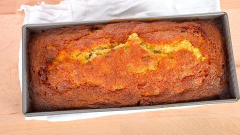 Ein goldbrauner, veganer Kuchen liegt in einer Kastenform auf einem hellen Holzuntergrund, umgeben von einem weißen Geschirrtuch.