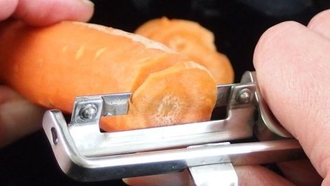 Eine karottenorange wird mit einem Sparschäler geschält, während sie über einen schwarzen Behälter gehalten wird.