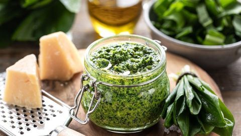 Ein Glas Bärlauchpesto steht auf einem Holzteller, umgeben von Öl, Käse und frischem Bärlauch im Hintergrund.