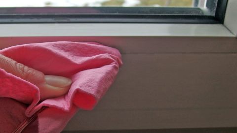 Ein pinkes Tuch reinigt schmale Ritzen am Fenster in einem hellen Raum mit Blick nach draußen.