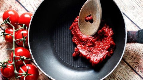 In einer Pfanne wird Tomatenmark mit einem Holzlöffel verrührt, während frische Tomaten am Rand liegen.