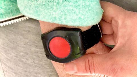 Ein roter Notrufknopf wird von einer Hand gedrückt, während er am Handgelenk einer Person befestigt ist, um Sicherheit zu gewährleisten.