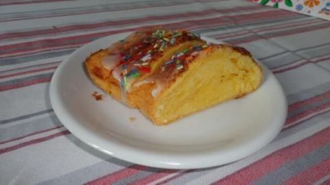 Ein Stück Napfkuchen mit Zuckerguss und bunten Streuseln liegt auf einem weißen Teller auf gestreifter Tischdecke.