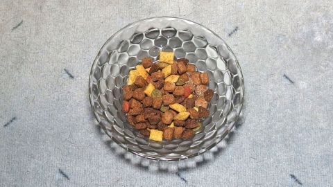 Eine Schüssel mit Trockenfutter und kleinen, gelben Stückchen steht auf einem hellen Untergrund, während eine Katze neugierig schaut.