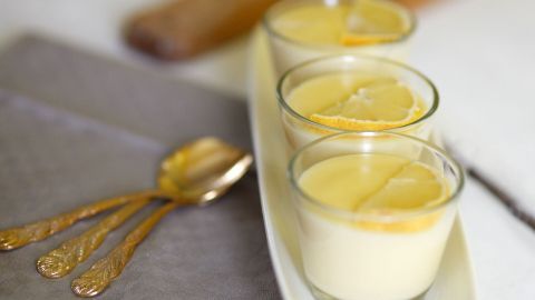 Drei Gläser mit Zitronen-Vanille-Creme stehen auf einer weißen Platte, umgeben von Besteck auf einem grauen Untergrund.