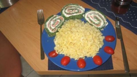 Eine bunte Portion Lachs-Frischkäse-Spinatrolle liegt auf einem blauen Teller, umgeben von Nudeln und Kirschtomaten.