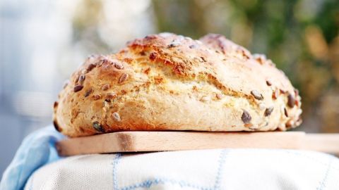 Ein frisch gebackenes Brot liegt auf einem Holzbrett, umgeben von einem karierten Tuch in einem hellen, einladenden Raum.