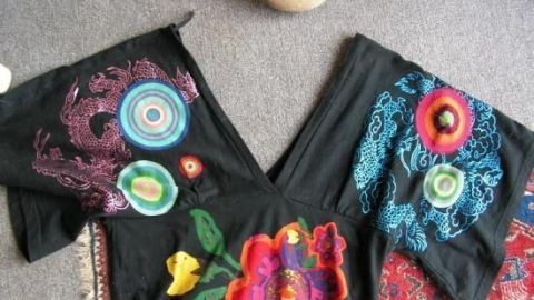 Ein schwarzes T-Shirt mit buntem, floralem Design liegt flach auf einem Teppich, während ein Stein daneben platziert ist.