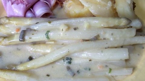 Weiße Spargelstangen liegen in einer cremigen Soße à la Hollandaise, angerichtet mit Schinken auf einem Teller.
