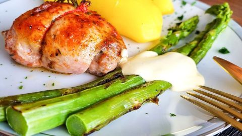 Auf dem Teller liegen zarte, grüne Spargelstangen mit Bechamel-Soße, untermalt von goldgelben Kartoffeln und einem gefüllten Fleischstück.