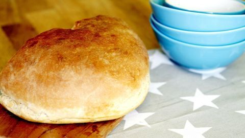Ein frisch gebackenes Brioche liegt auf einem Holzbrett, während bunte Schalen im Hintergrund bereitstehen.
