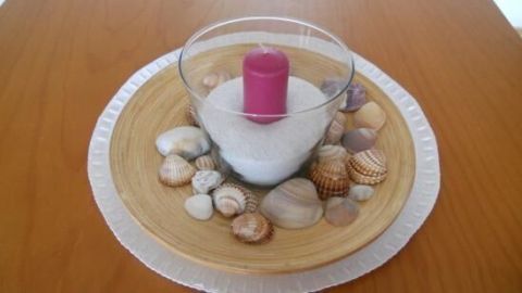 Eine pinke Kerze steht in einem Glas auf Sand, umgeben von dekorativen Muscheln auf einem Holztablett.