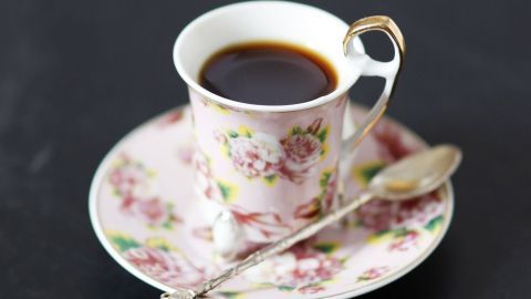 Eine elegante Tasse Schwarztee steht auf einer Blumenteller, daneben liegt ein Löffel aus Metall.