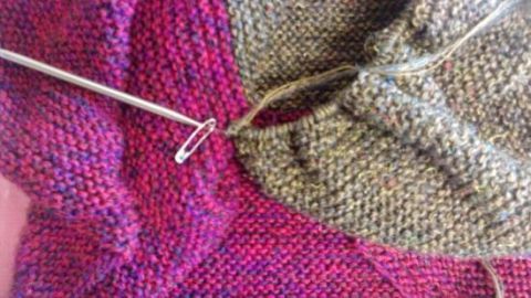 Ein Strickstück in bunten Farben liegt daneben, während ein Maschenstopper seine Funktion im Gewebe erfährt.