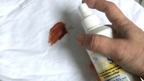 Ein Hand Spray entfernt einen roten Marinadefleck von einem weißen Kleidungsstück auf einem Holzuntergrund.