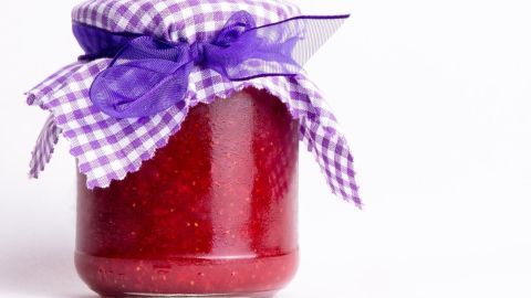 Ein Glas mit roter Marmelade steht auf weißem Hintergrund, bedeckt mit einem lilafarbenen karrierten Stoff und Schleife.