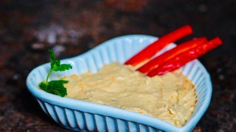 Ein Herzschälchen mit Hummus Dip wird präsentiert, garniert mit frischen Paprikastreifen und einem Blatt Koriander.