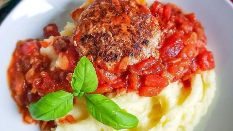 Ein saftiger Frikadelle liegt auf einem Bett aus cremigem Kartoffelpüree, garniert mit würziger Tomatensauce und frischem Basilikum.