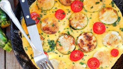 Ein Omelette, belegt mit Zucchini und Tomaten, liegt auf einem schwarzen Teller, während Besteck daneben bereitliegt.